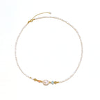 Delicate white beaded necklace with baroque pearl center, gold accents, and coral and aqua gemstones on cream background.
